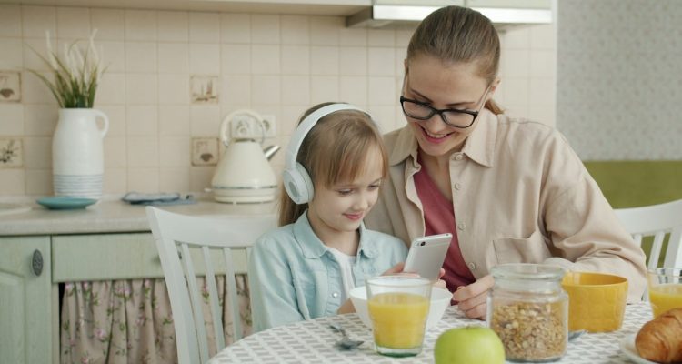 Slow motion of happy little girl and loving mother using smartphone and listening to music in headphones at kitchen table. Childhood and motherhood concept.